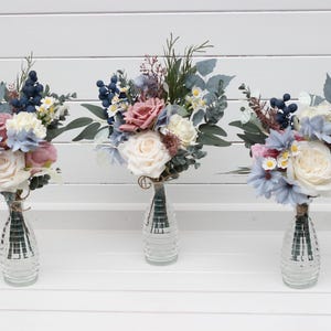 May include: Three flower bouquets in clear glass vases. The bouquets have light blue, pink, and cream flowers, with dark blue berries and green leaves. The vases are clear with a ribbed design.