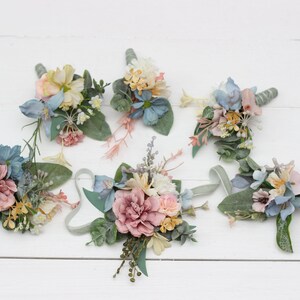 May include: Six small bouquets of artificial flowers in shades of pink, blue, and cream. The bouquets are arranged on a white wooden surface.