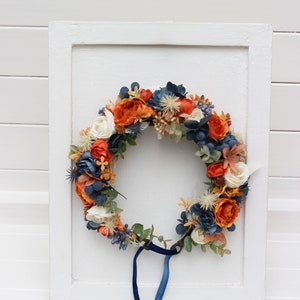 May include: A floral wreath with orange, white, and blue flowers, with a blue velvet ribbon attached.