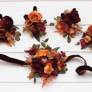 May include: A collection of artificial flowers in shades of burgundy, orange, and gold, including roses, arranged as boutonnieres and a wrist corsage. The boutonnieres have black stems, and the wrist corsage has a dark brown velvet ribbon.