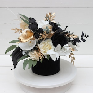 May include: A floral arrangement in a black velvet container. The bouquet features white, black, and cream-colored flowers with gold accents and green leaves. The arrangement is displayed on a white, round pedestal.
