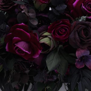 May include: A bouquet of deep purple and burgundy roses, with green leaves and other flowers, arranged in a lush, romantic style.