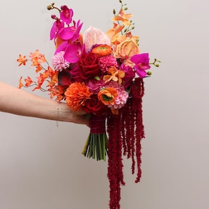 May include: A colourful bouquet featuring red roses, pink orchids, and orange blooms, with trailing red amaranthus. The flowers are held in a hand against a plain backdrop, showcasing a variety of textures and hues.
