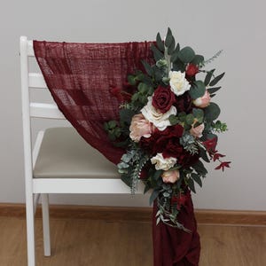 May include: A white chair decorated with a burgundy fabric sash and a bouquet of burgundy, blush, and white flowers with greenery.