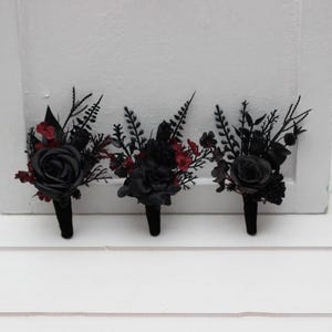 May include: Three black boutonnieres with dark red accents. Each boutonniere features a black rose and various dark green and red foliage. The stems are wrapped in black fabric. Ideal for a wedding or formal event.