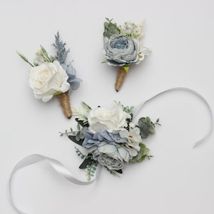 May include: Three floral arrangements with white and blue flowers. Two are boutonnieres with twine-wrapped stems. The third is a wrist corsage with a white ribbon. The flowers include roses and other greenery.