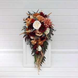 May include: A cascading bridal bouquet featuring a mix of burgundy, peach, and white flowers. The bouquet is crafted with silk flowers and greenery, showcasing a variety of textures and shapes.