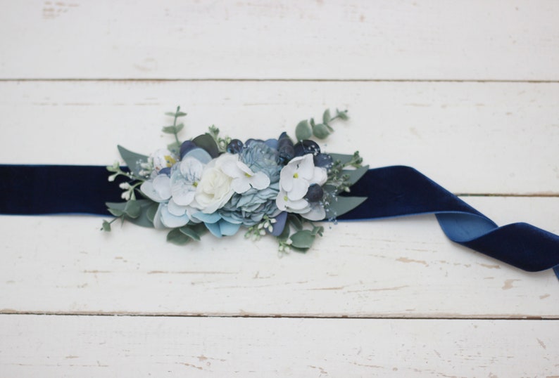 Pu&ograve; includere: Un nastro di velluto blu navy con un abbellimento floreale. L'abbellimento presenta fiori bianchi, blu e verdi.