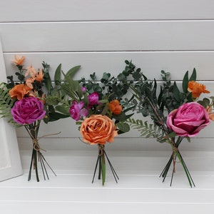 May include: Three bouquets of artificial flowers, each with a different colour scheme. The first bouquet is pink and orange, the second is orange and pink, and the third is pink and orange. The bouquets are arranged on a white surface.