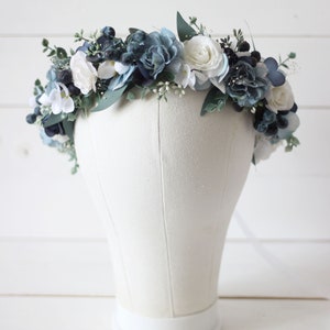 Pu&ograve; includere: Una corona floreale blu e bianca con rose bianche e fiori blu. La corona &egrave; su una testa di manichino bianca.