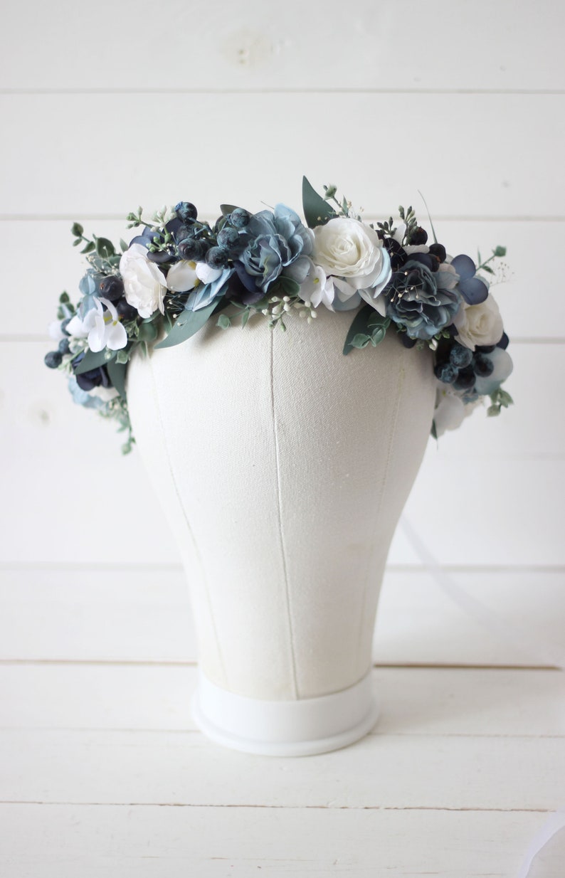 Pu&ograve; includere: Una corona floreale blu e bianca con rose bianche e fiori blu. La corona &egrave; su una testa di manichino bianca.
