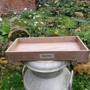 May include: A rectangular wooden tray with a light brown finish. The tray has a small metal plate with the text "Hop Inn" on the front. The tray is sitting on top of a large, silver metal container, outdoors.