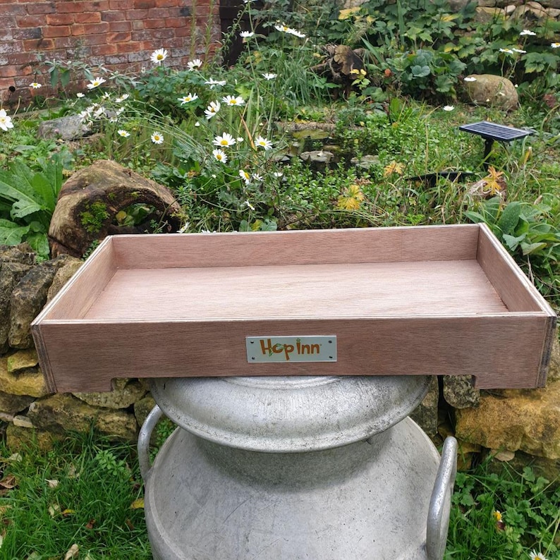 May include: A rectangular wooden tray with a light brown finish. The tray has a small metal plate with the text "Hop Inn" on the front. The tray is sitting on top of a large, silver metal container, outdoors.