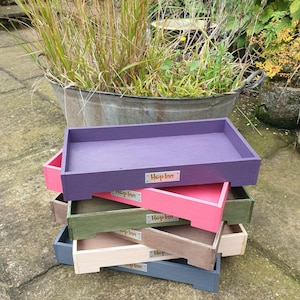 May include: A stack of rectangular wooden trays in various colors: purple, pink, green, beige, and gray. Each tray has a small rectangular label that reads "Hop Inn". The trays are stacked on top of each other.