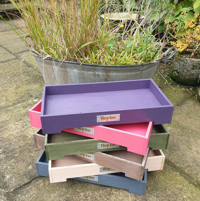 May include: A stack of rectangular wooden trays in various colors: purple, pink, green, beige, and gray. Each tray has a small rectangular label that reads "Hop Inn". The trays are stacked on top of each other.