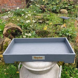 May include: A rectangular, gray wooden tray with the text "Hop Inn" on a small metal plate. The tray is sitting on top of a large, silver metal container, with a garden in the background.