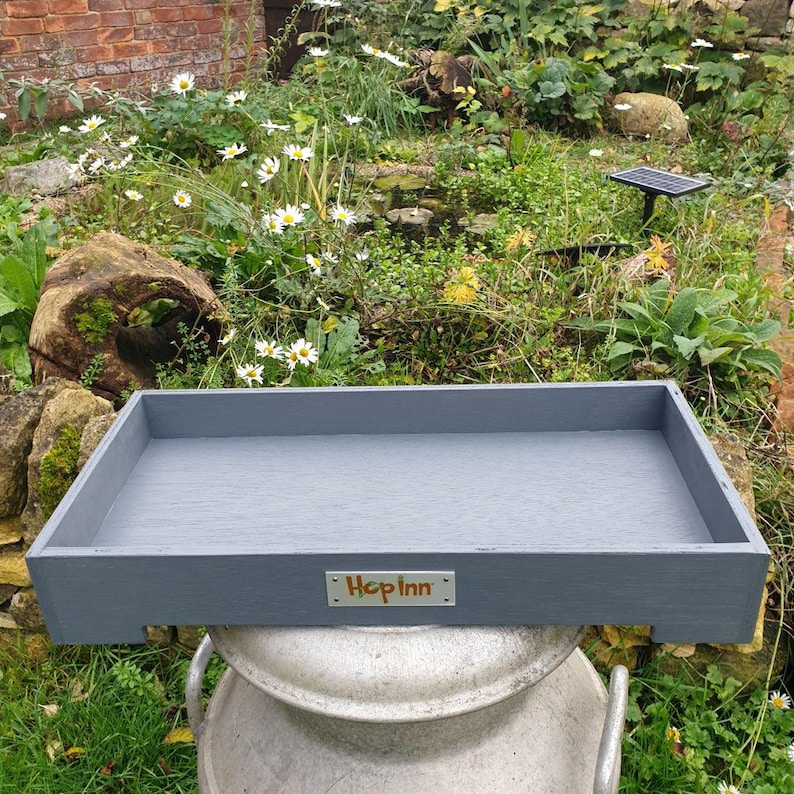 May include: A rectangular, gray wooden tray with the text "Hop Inn" on a small metal plate. The tray is sitting on top of a large, silver metal container, with a garden in the background.