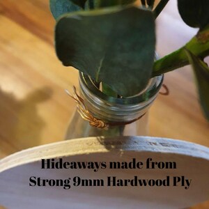 Op de afbeelding: Een heldere glazen fles met een plantenstengel, vastgezet met gouddraad. De fles staat op een licht houten voet. Tekst op de basis luidt: "Hideaways made from Strong 9mm Hardwood Ply."