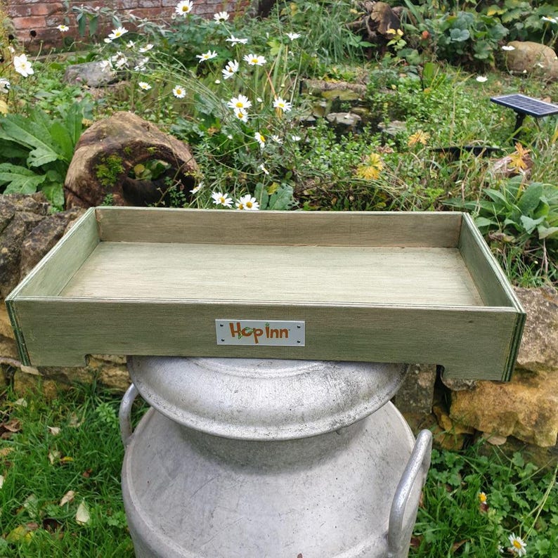 May include: A rectangular wooden tray with a light green finish, resting on a metal container. The tray has a simple design with a raised edge and the text "Hop Inn" on a small metal plate. The background features greenery and flowers.