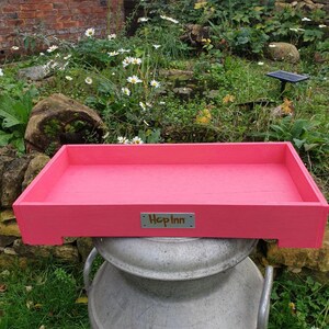 May include: A rectangular, pink wooden tray with the text "Hop Inn" on a small metal plate. The tray is resting on a large, silver metal container. The background features greenery, flowers, and a brick wall.
