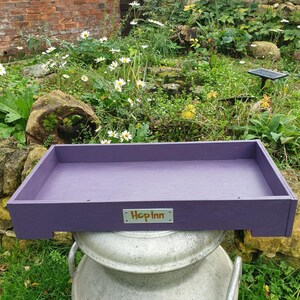 May include: A rectangular, purple wooden tray with the text "Hop Inn" on a small metal plate. The tray is placed on top of a large, silver metal container, with a garden in the background.