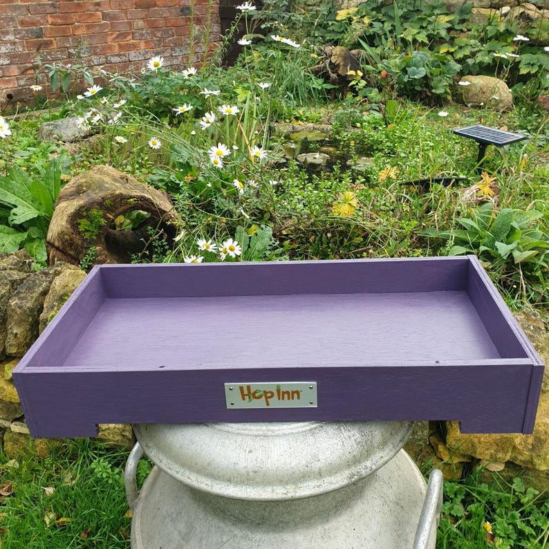 May include: A rectangular, purple wooden tray with the text "Hop Inn" on a small metal plate. The tray is placed on top of a large, silver metal container, with a garden in the background.