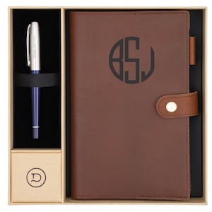 Puede incluir: Un juego de regalo que incluye un diario de cuero marrón con un monograma y un bolígrafo a juego. El diario tiene un cierre de botón y un portalápices. El bolígrafo es azul y plateado, con el texto "Blake Stevens".