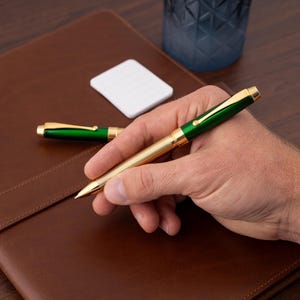 Puede incluir: Un bol&iacute;grafo dorado y verde con un ca&ntilde;&oacute;n texturizado. El bol&iacute;grafo est&aacute; en una mano, y hay un cuaderno de cuero marr&oacute;n en el fondo.