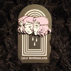 May include: A black and white enamel pin featuring a cartoon image of a woman's face with pink hair. The pin is set against a black background with a white border and the text "LULU VANHOAGLAND".