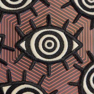 May include: Embroidered patches with a stylized eye design. The patches feature concentric white and black circles, black eyelashes, and are set against a geometric background with red and grey stripes.