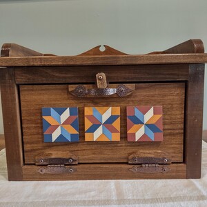 May include: Wooden bread box with a dark stain. The box features three square, colorful star quilt block designs. The box has a hinged door with a copper-colored latch and hinges. The top of the box has a curved design.