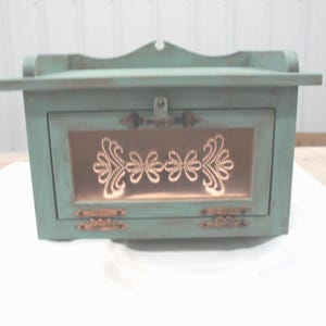 May include: A teal-colored wooden box with a decorative copper-colored floral design on the front. The box has a hinged door, a small handle, and decorative hardware. The top of the box has a curved design.