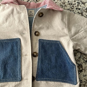 May include: A beige jacket with two large blue denim pockets and a red and white striped collar. The jacket has brown buttons and a light blue lining. The tag reads "TAYLOR MADE".