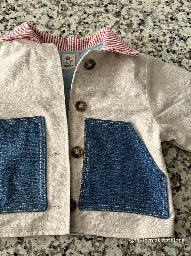 May include: A beige jacket with two large blue denim pockets and a red and white striped collar. The jacket has brown buttons and a light blue lining. The tag reads "TAYLOR MADE".