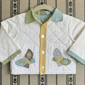 May include: A white quilted jacket with butterfly appliqu&eacute;s. The jacket has a yellow centre, green and blue accents, and a diamond pattern. It features two pockets with butterfly designs and is buttoned up.