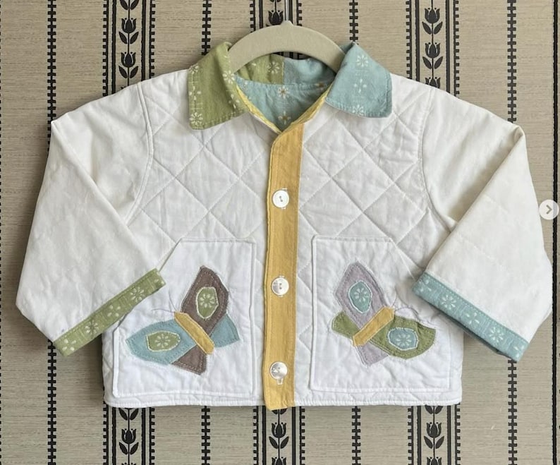 May include: A white quilted jacket with butterfly appliqu&eacute;s. The jacket has a yellow centre, green and blue accents, and a diamond pattern. It features two pockets with butterfly designs and is buttoned up.