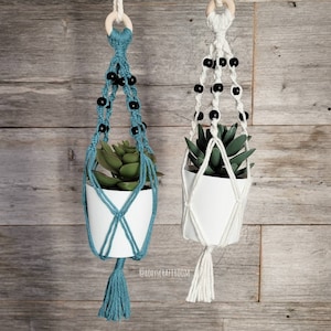 Op de afbeelding: Twee macramé plantenhangers met zwarte kralen en houten ringen. Eén is teal en de andere is wit. Beide houden kleine witte potten met groene planten.