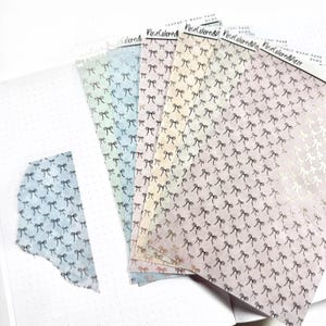 Op de afbeelding: Een set van zes pastelkleurige washi tape rollen met een herhalend patroon van zwarte strikken. De washi tape is gelabeld "RoseColoredBlaze" en is verpakt in doorzichtig plastic.