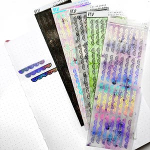 May include: A set of six holographic sticker sheets featuring a scalloped edge design with a spiderweb pattern. The stickers are in various colours, including purple, blue, green, and yellow.