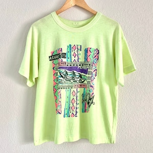 Pode incluir: Uma camiseta verde claro com um design geométrico colorido e o texto "PRIMITIVE" na frente.
