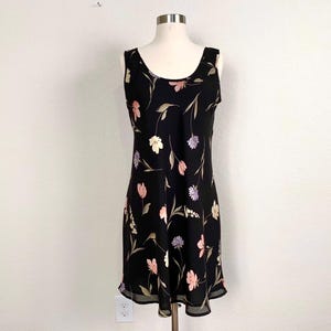 May include: A black sleeveless dress with a floral pattern in pink, white, and purple. The dress has a scoop neckline and a slightly flared skirt. The dress is displayed on a mannequin against a white wall.