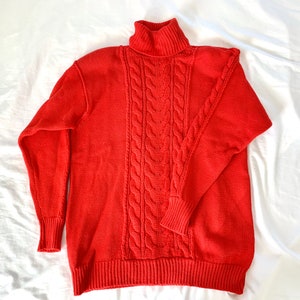 Könnte beinhalten: Ein roter Pullover mit Zopfmuster und Rollkragen. Der Pullover hat ein klassisches Zopfmuster und einen hohen Rollkragen.