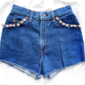 May include: Blue denim shorts with a frayed hem and pink floral trim around the pockets.