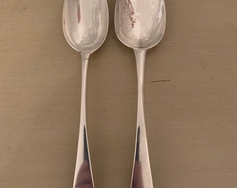 May include: Two silver spoons with ornate handles. The spoons have engraved initials on the back of the handles. The spoons are on a light brown surface.