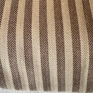 K&ouml;nnte beinhalten: Nahaufnahme eines strukturierten Stoffes mit einem sich wiederholenden vertikalen Streifenmuster. Die Streifen wechseln sich zwischen hellem Beige und Dunkelbraun ab, mit einem subtilen Fischgr&auml;tgewebe. Der Stoff scheint ein gewebtes Textil zu sein.