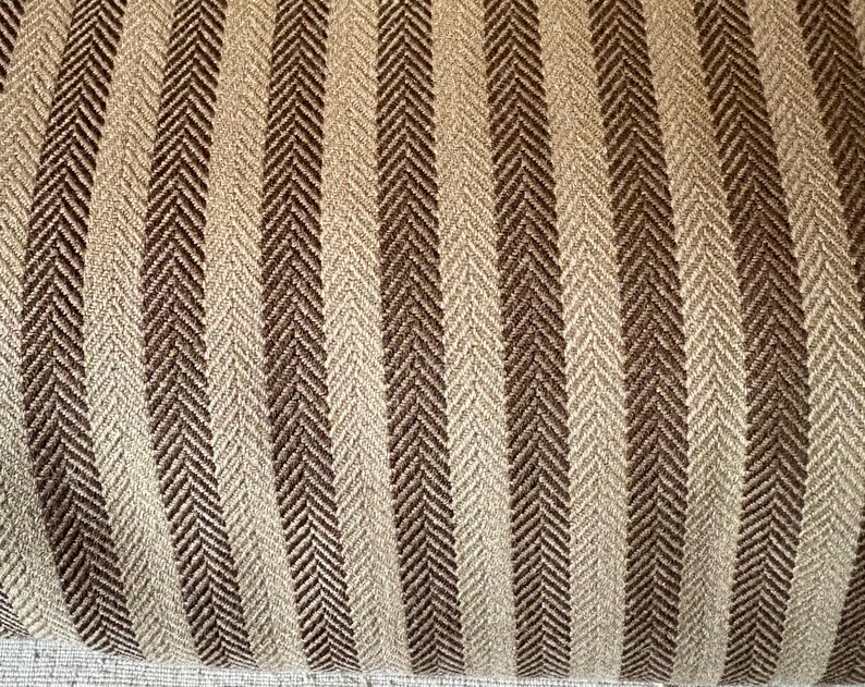 K&ouml;nnte beinhalten: Nahaufnahme eines strukturierten Stoffes mit einem sich wiederholenden vertikalen Streifenmuster. Die Streifen wechseln sich zwischen hellem Beige und Dunkelbraun ab, mit einem subtilen Fischgr&auml;tgewebe. Der Stoff scheint ein gewebtes Textil zu sein.