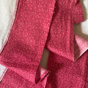 Op de afbeelding: Close-up van verschillende stroken rode stof met een klein wit stippenpatroon. De stof is gevouwen en gedrapeerd, waardoor de achterkant van de stof zichtbaar is. De stof lijkt katoen of een vergelijkbaar materiaal te zijn.