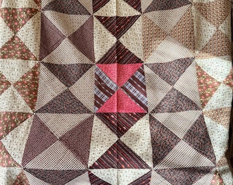 May include: A patchwork quilt with a geometric design. The quilt features triangles and squares in shades of brown, cream, and pink. The pattern includes floral and plaid fabrics. The edges are trimmed with a solid pink border.