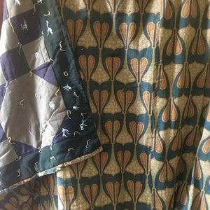 Puede incluir: Primer plano de una colcha con un diseño de patchwork en tonos grises, negros y blancos. La colcha está doblada sobre una tela con un patrón repetitivo de formas de lágrima marrones sobre un fondo azul.