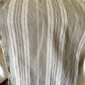 Puede incluir: Primer plano de un trozo de tela con rayas verticales en tonos grises y blancos. La tela tiene un aspecto texturizado y bordes deshilachados, lo que sugiere un artículo hecho a mano o vintage. Las rayas varían en ancho.